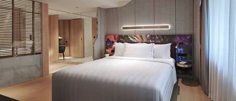 A modern hotel room with a large bed, a headboard with a floral design, a bedside table with a lamp, and a view of the city through large windows.