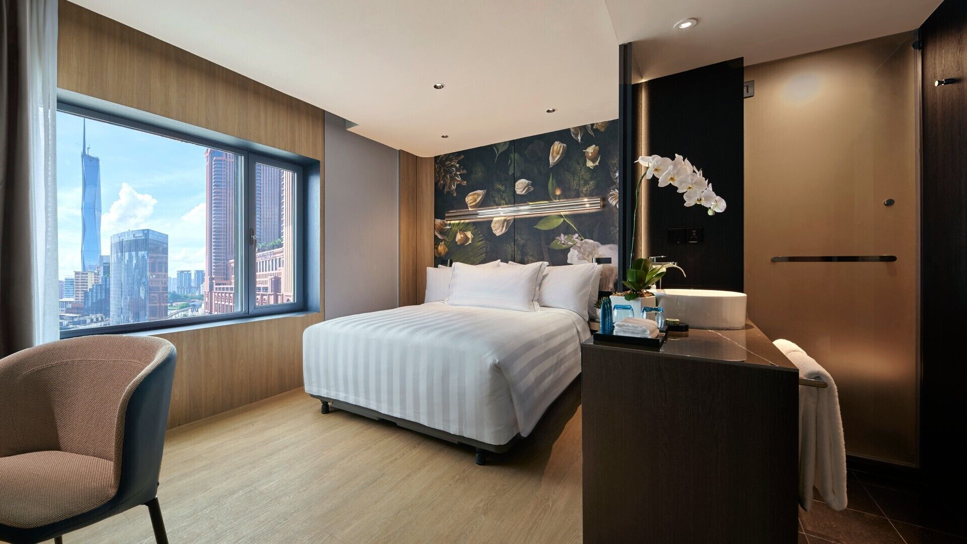 A modern hotel room with a large bed, a desk with a sink, a chair, and a view of the city skyline.