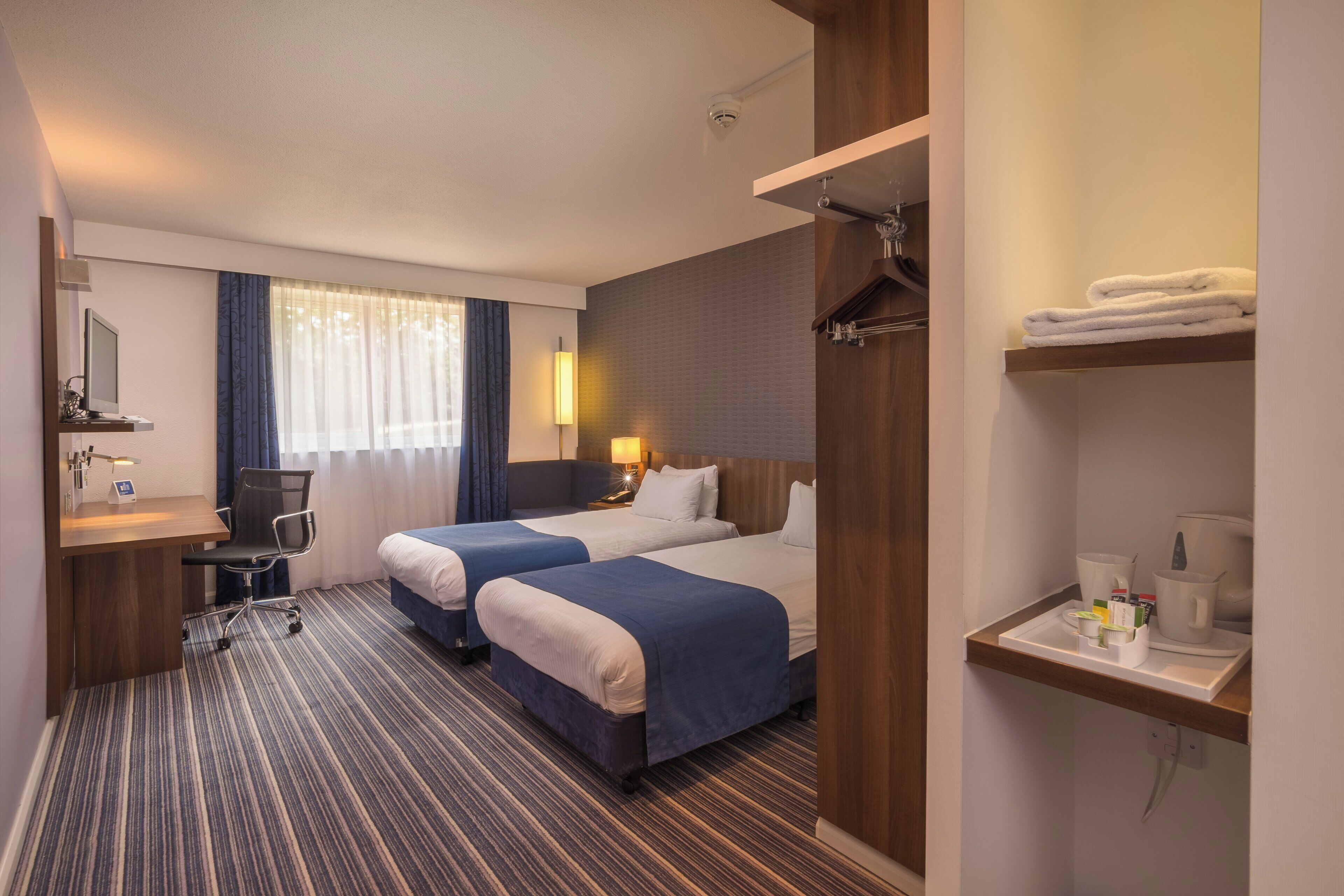 Habitación de hotel con dos camas, un escritorio, una silla, lámpara de escritorio, computadora, cafetera y estante con toallas.