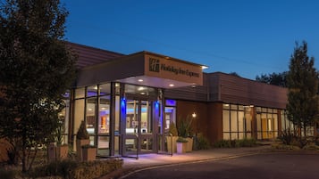 Un hotel Holiday Inn Express con entrada de vidrio, asientos al aire libre y fachada bien iluminada.