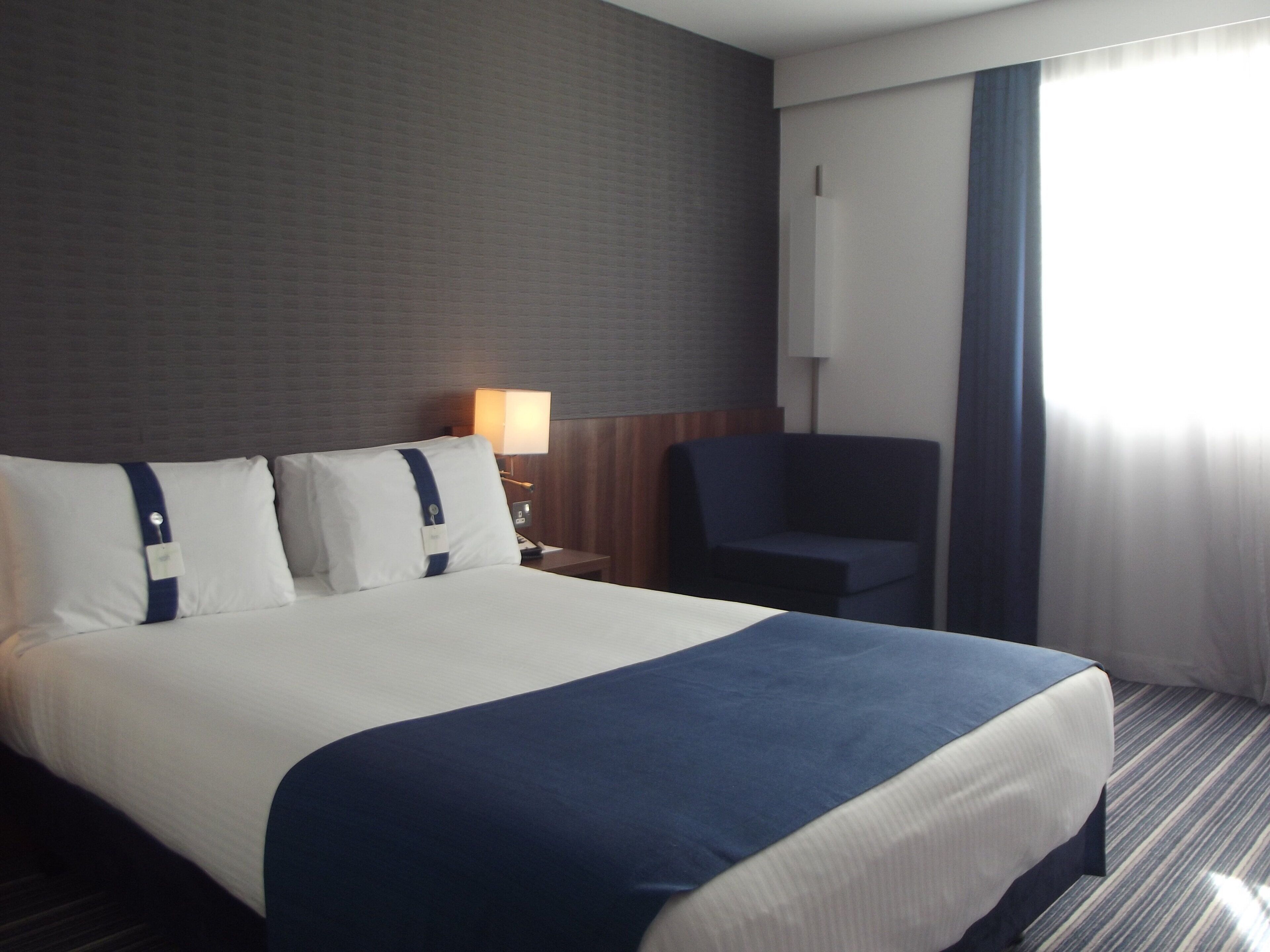 Una camera d'albergo con un letto grande, un comodino con una lampada, una sedia e una finestra con tende.