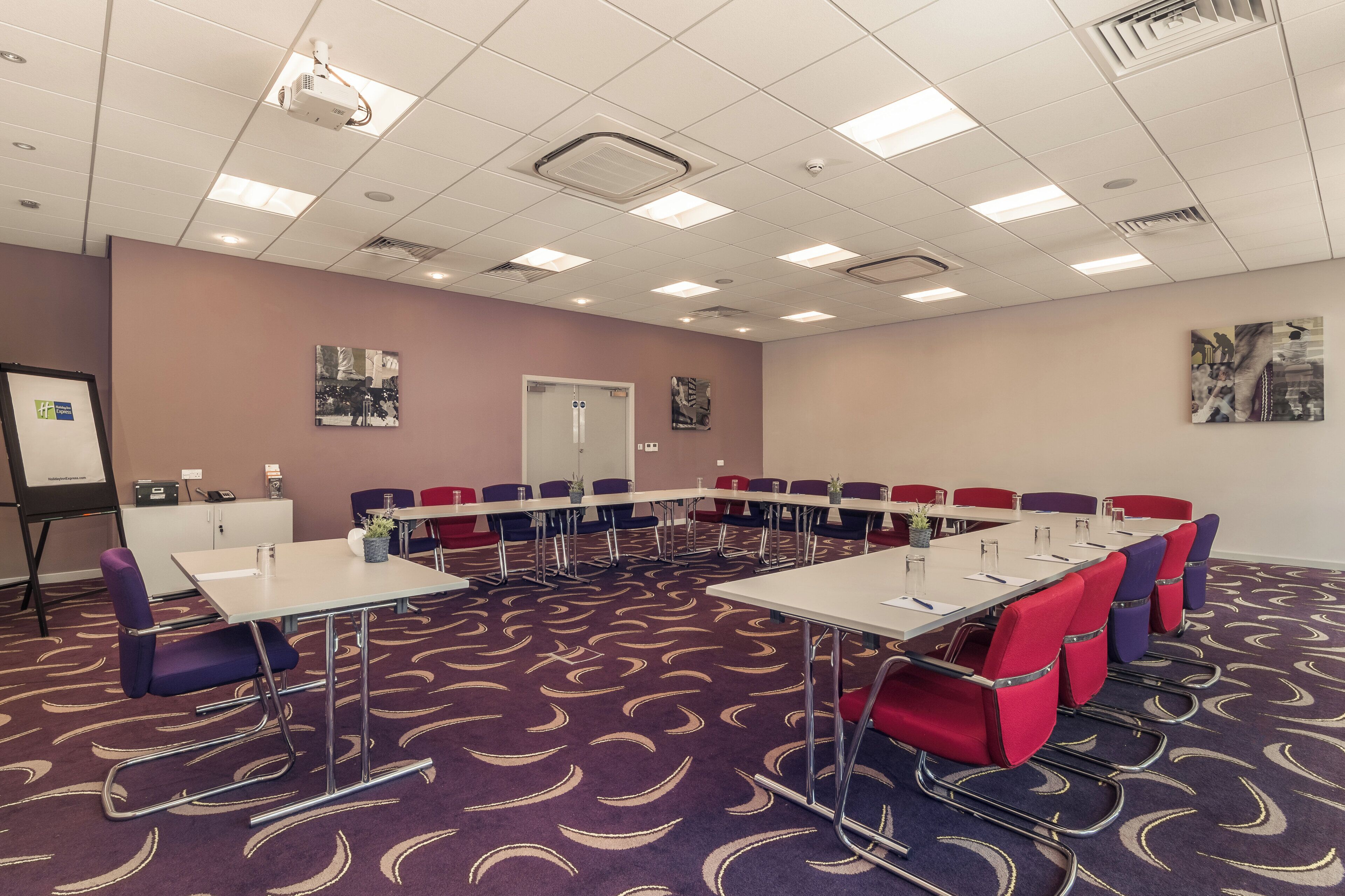 A conference room with a long table, multiple chairs, a whiteboard, and a projector on the ceiling.