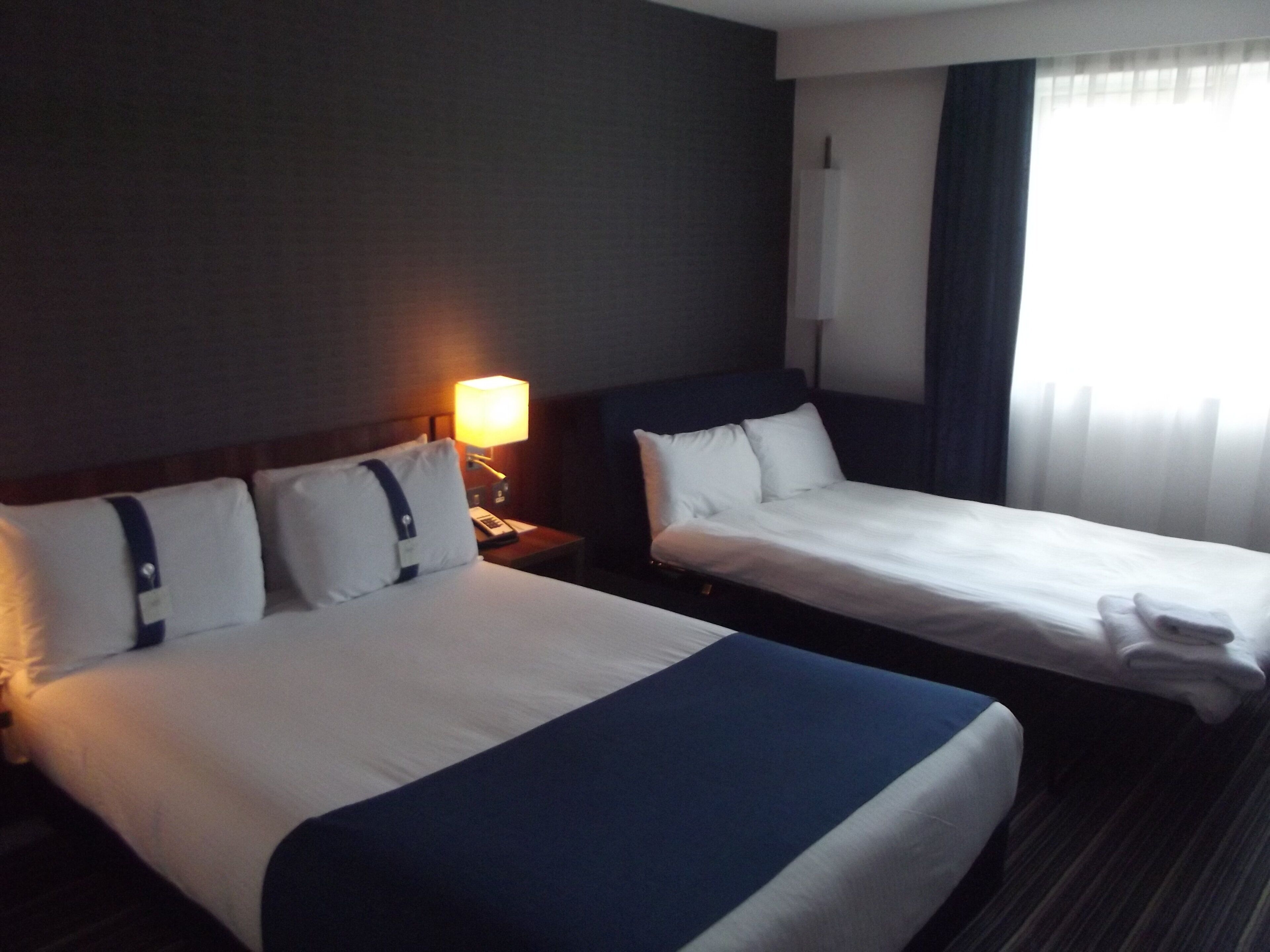 A hotel room with two beds, a nightstand with a lamp, and a window with curtains.