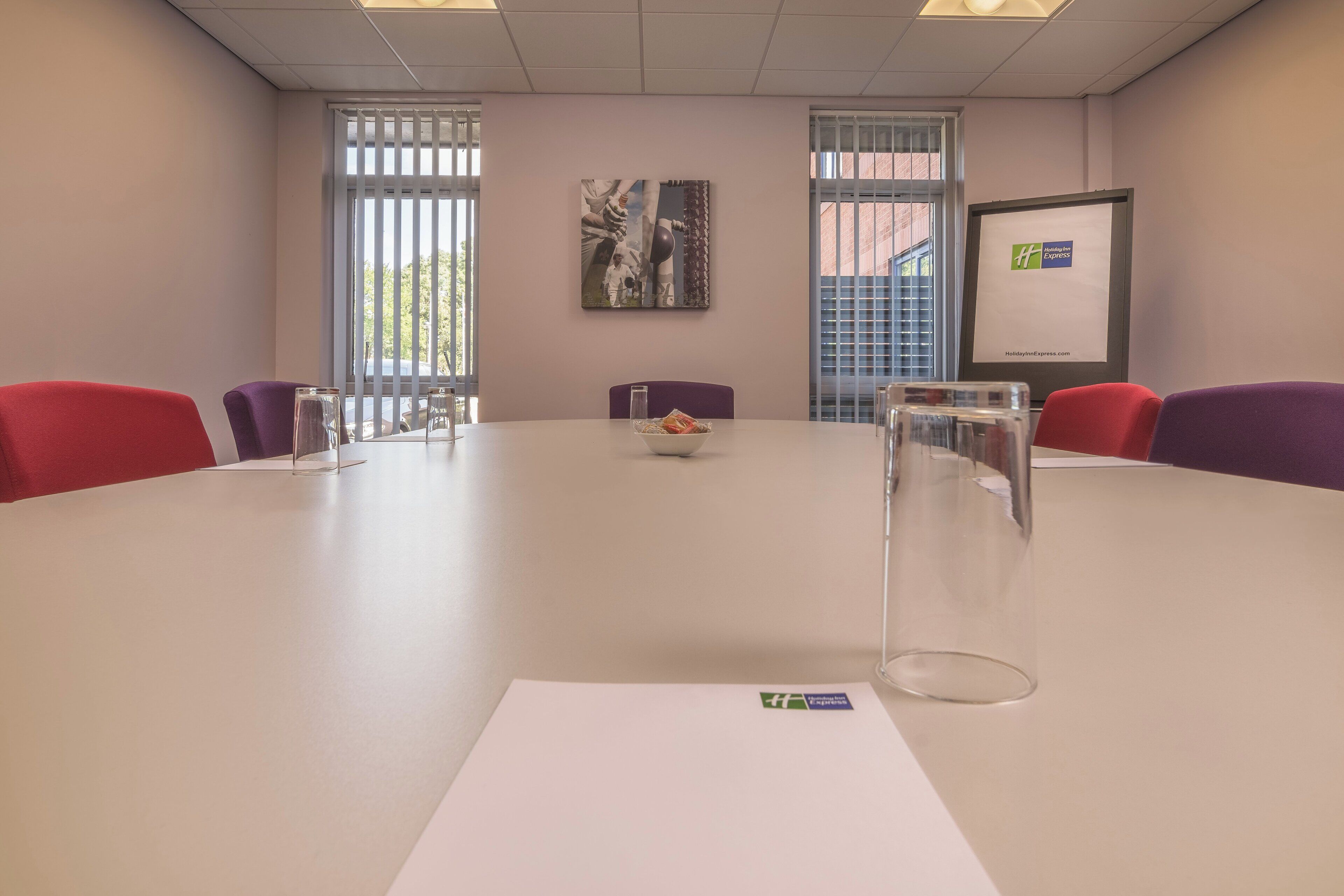 A conference room with a large table, red chairs, a whiteboard, and a framed picture on the wall.