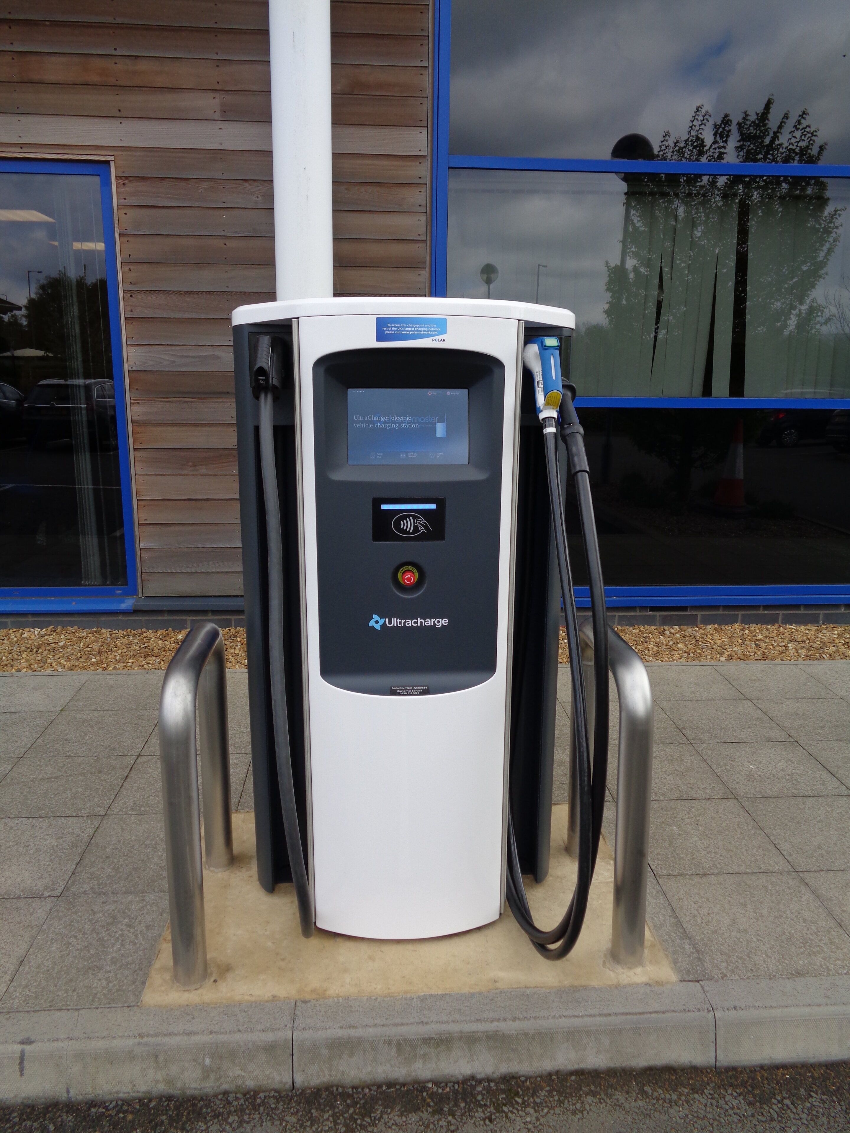 Una estación de carga para vehículos eléctricos con pantalla digital, marca Ultracharge, ubicada frente a un edificio con revestimiento de madera y ventanas con marco azul.