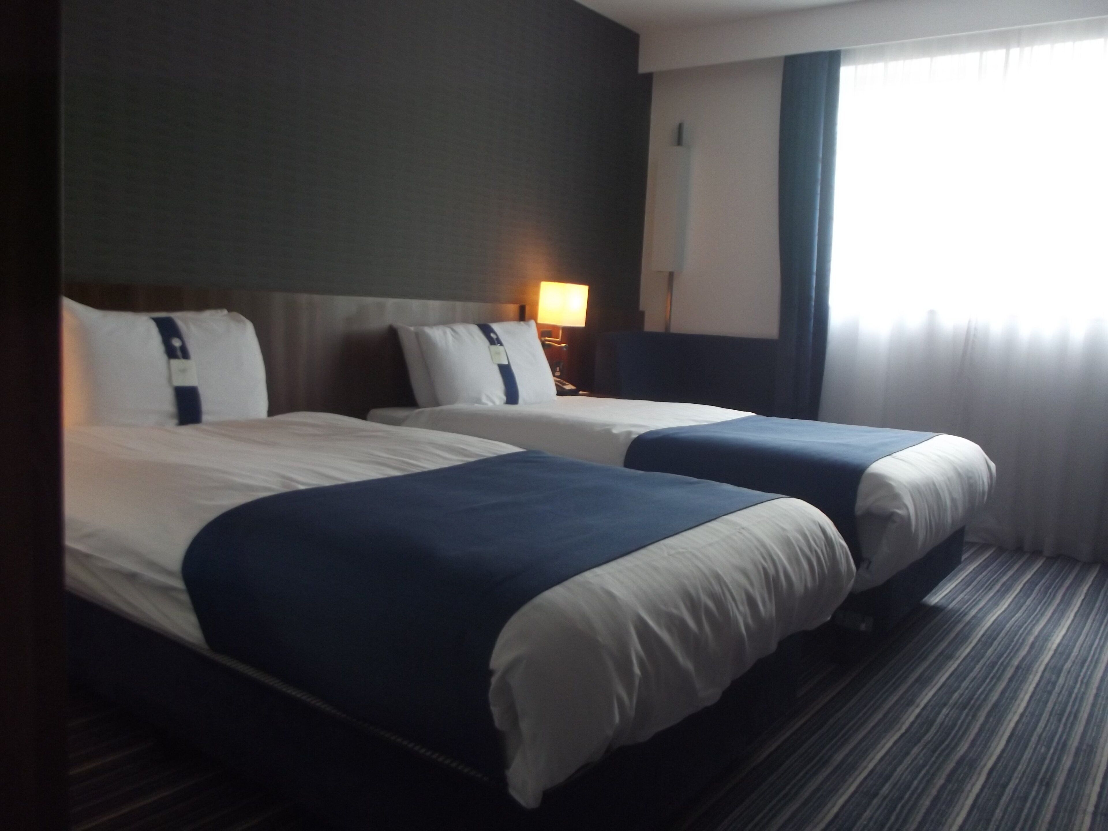 A hotel room with two beds, a nightstand, a lamp, and a window with curtains.