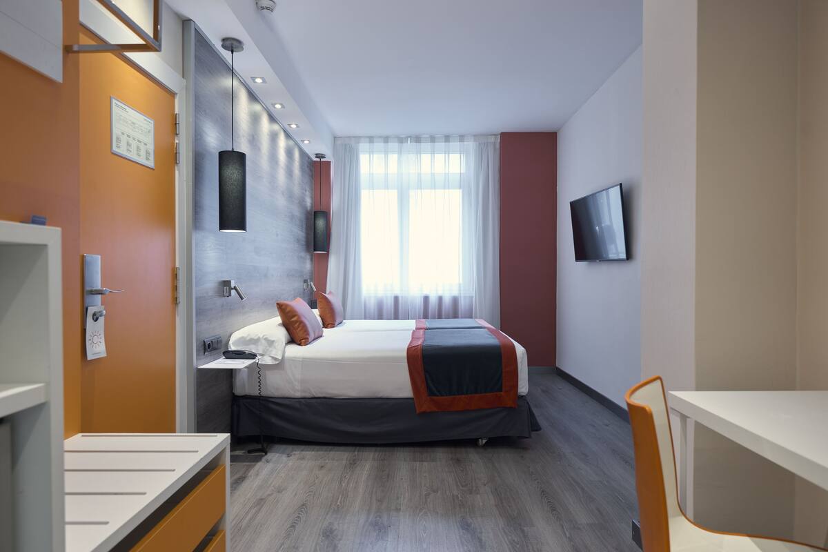 A modern hotel room with a bed, bedside table, and a TV mounted on the wall.