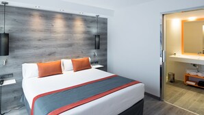 A modern hotel room with a large bed, a wooden headboard, a bedside table with a lamp, and a bathroom visible through an open door. - Catalonia Mikado (Barcelona)