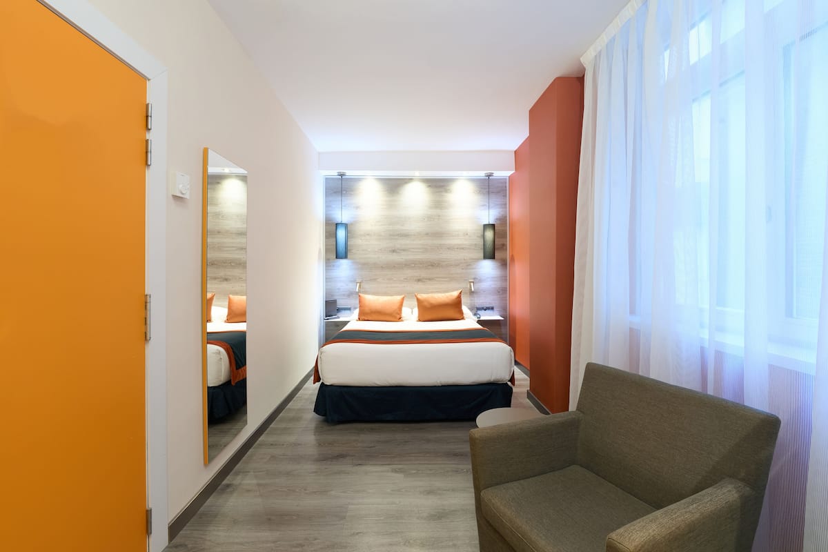 A hotel room with a bed, a chair, a mirror, and a window with curtains.
