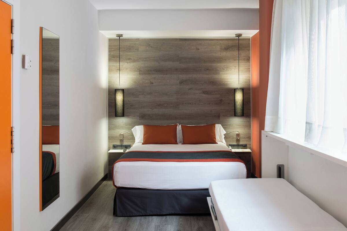 A modern hotel room with a large bed, a smaller bed, a wooden accent wall, and a window with sheer curtains.