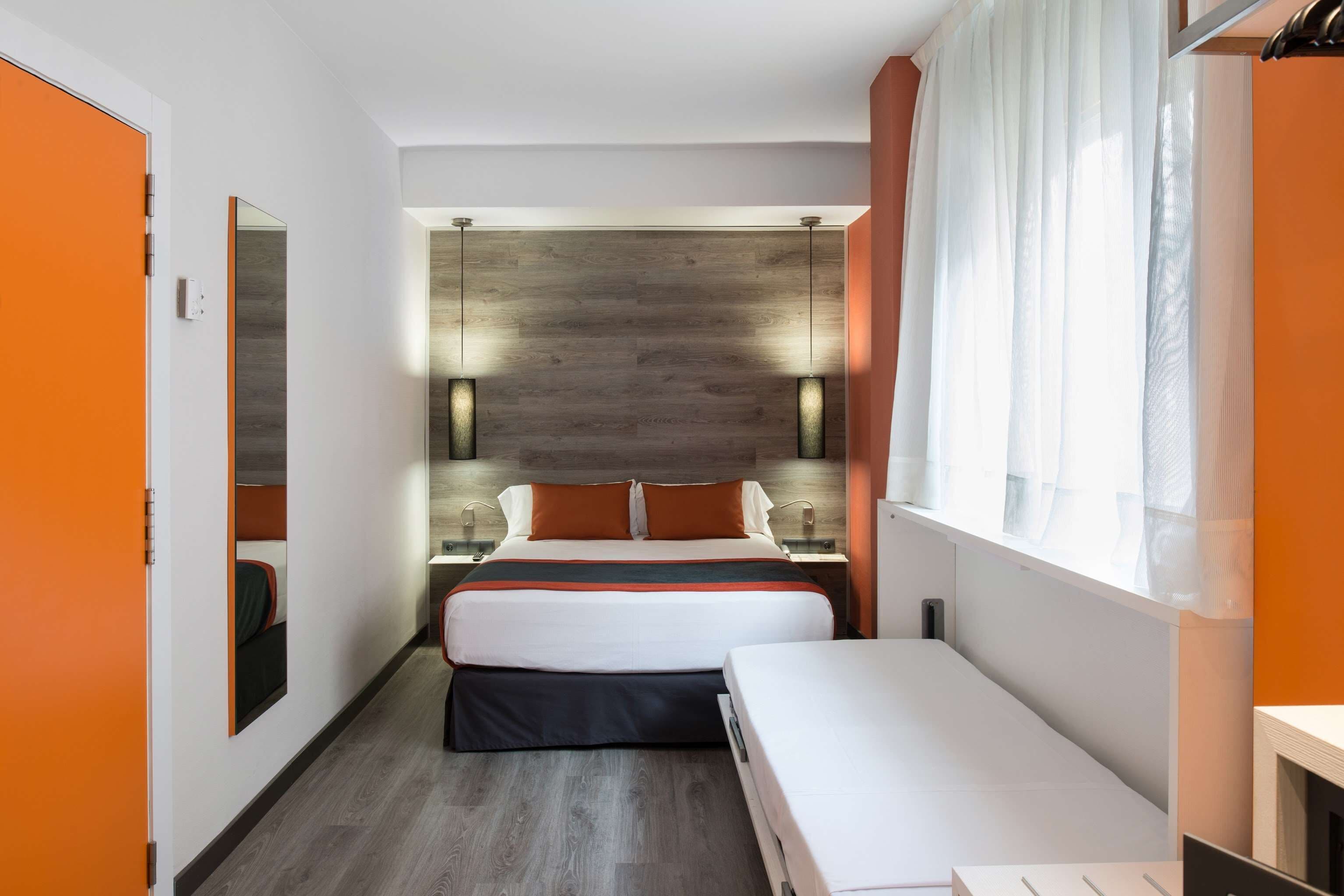 A modern hotel room with a large bed, a smaller bed, a nightstand, and a window with sheer curtains.