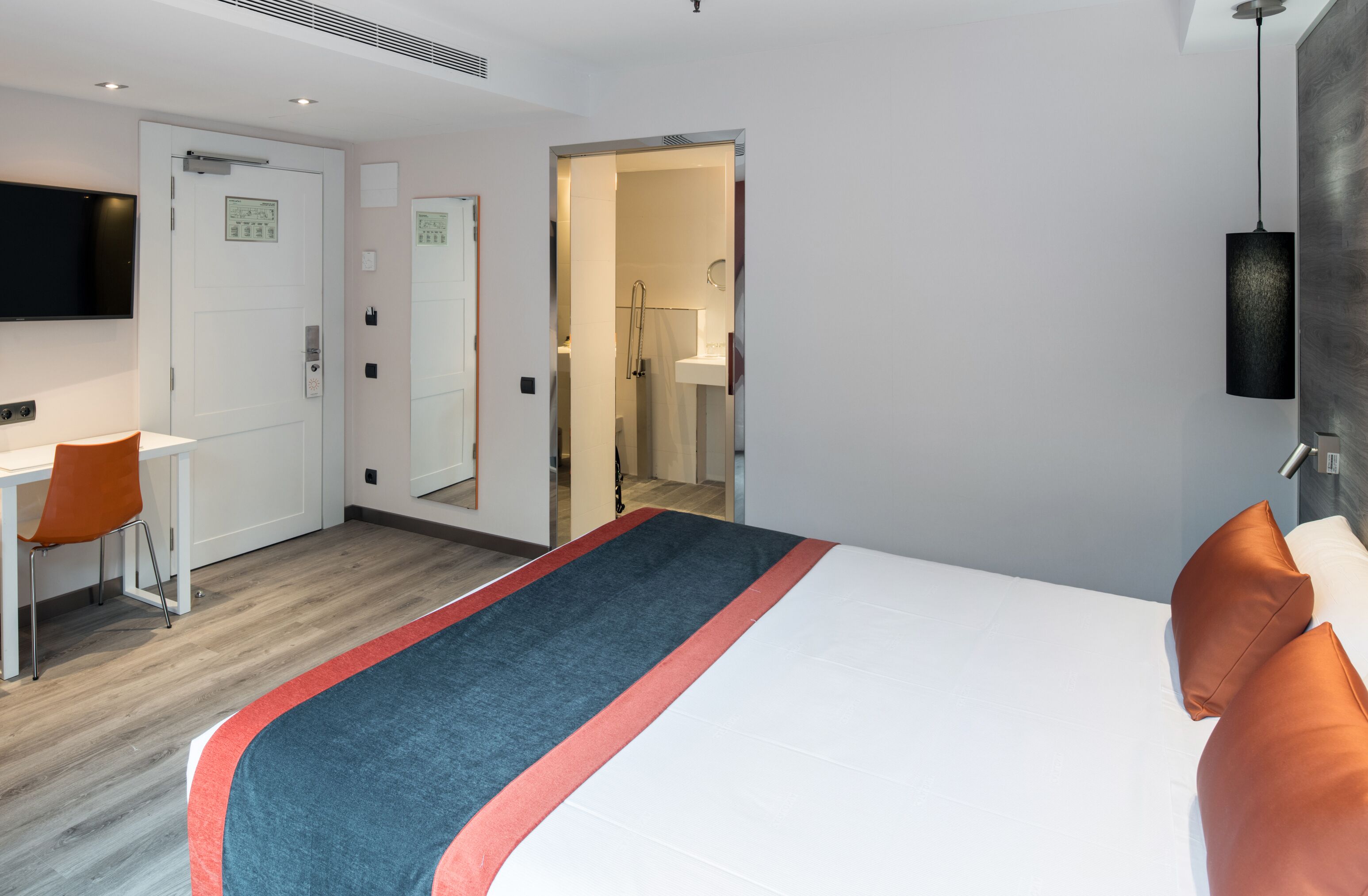 A hotel room with a bed, a desk, a chair, a TV, and a bathroom visible through an open door.