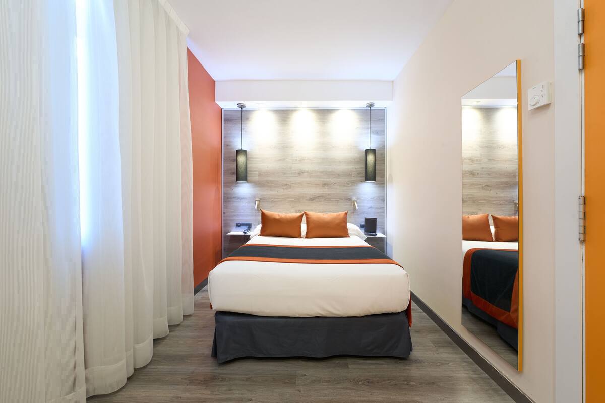 A modern hotel room with a large bed, a nightstand, a mirror, and a view of the hallway.