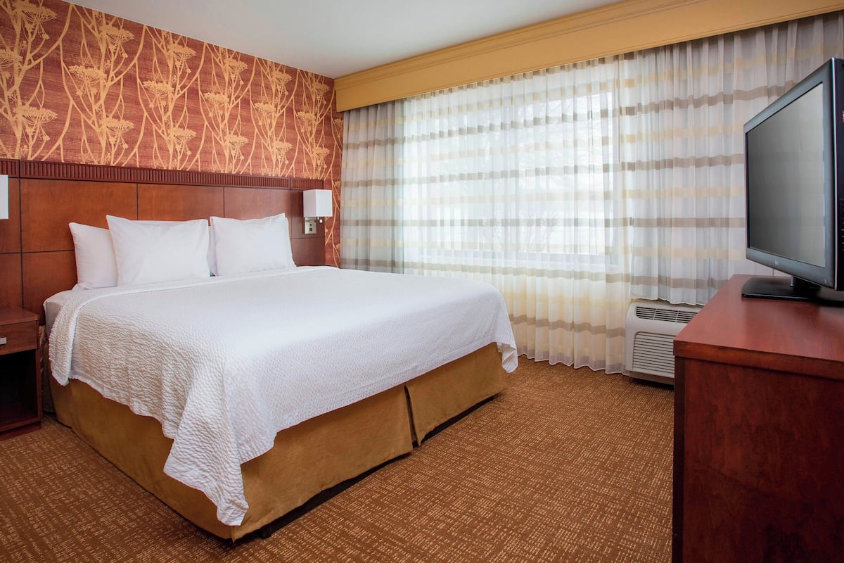 Habitación de hotel con una cama grande, cabecera de madera, televisor de pantalla plana y una ventana con cortinas translúcidas.