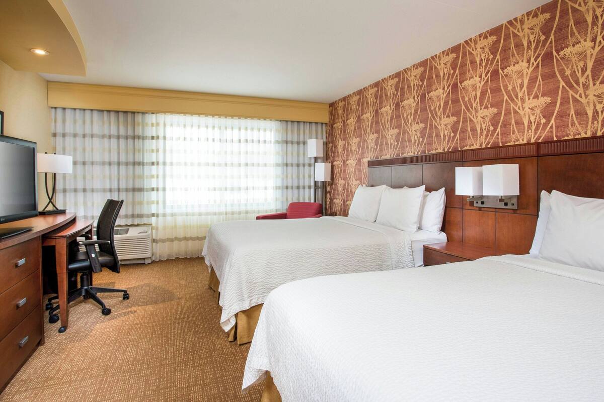 Habitación de hotel con dos camas, un escritorio con silla, televisor y un ventanal con cortinas.