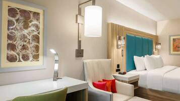 A hotel room with a desk, a chair, a bed with a blue headboard, and a wall-mounted lamp.