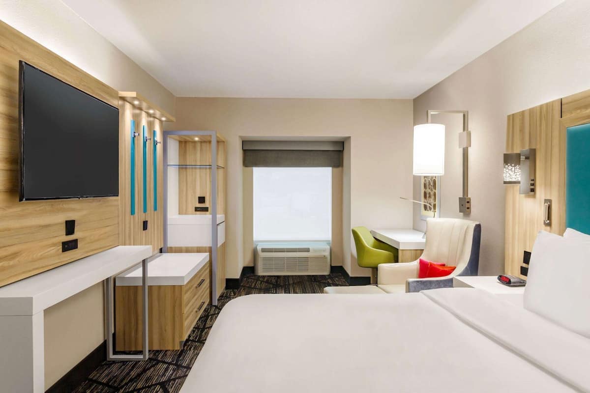 A hotel room with a large bed, a desk with a chair, a television, a window, and a wall-mounted lamp.
