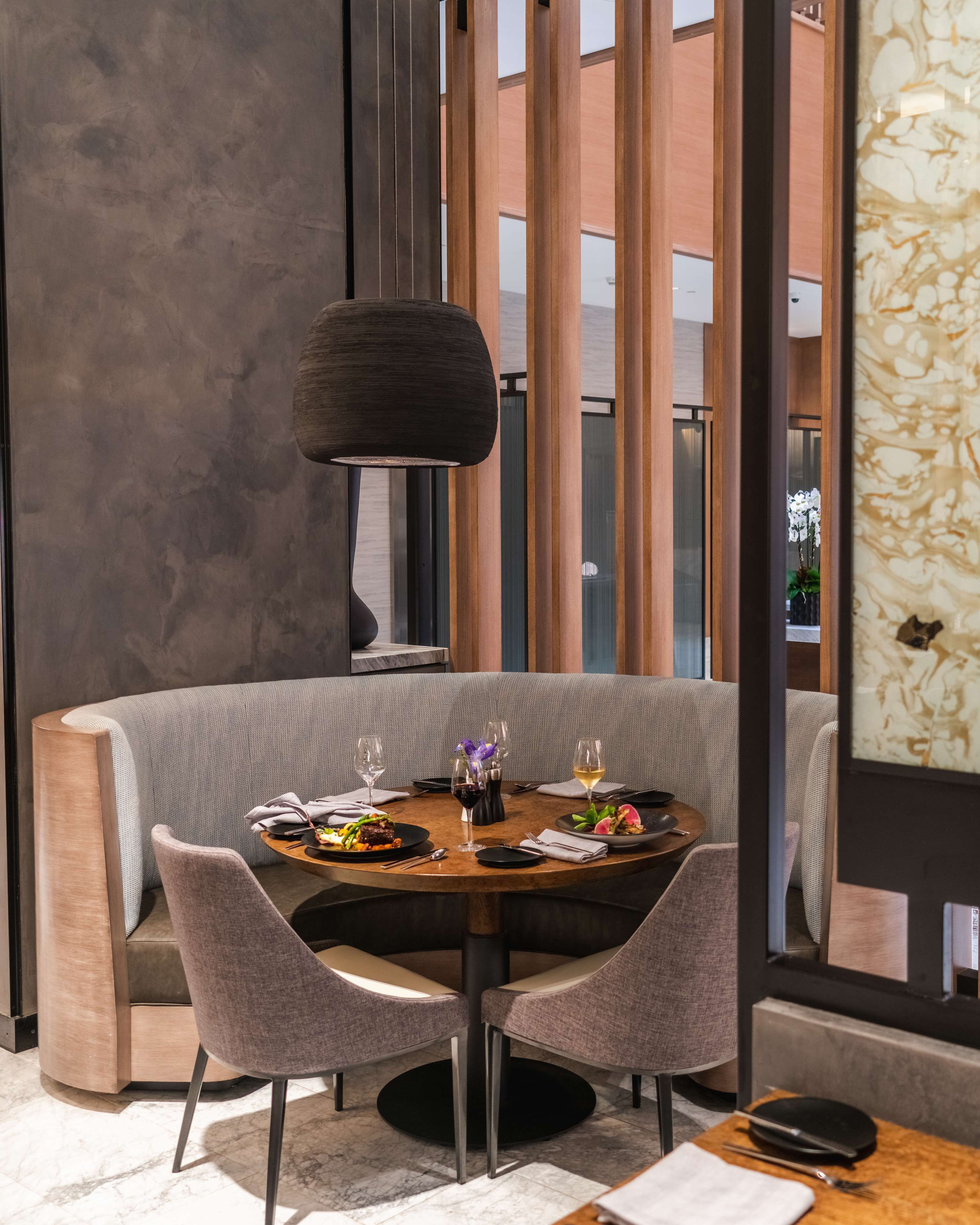 A modern dining area with a round table, two chairs, a lamp, and a view of the city through large windows.