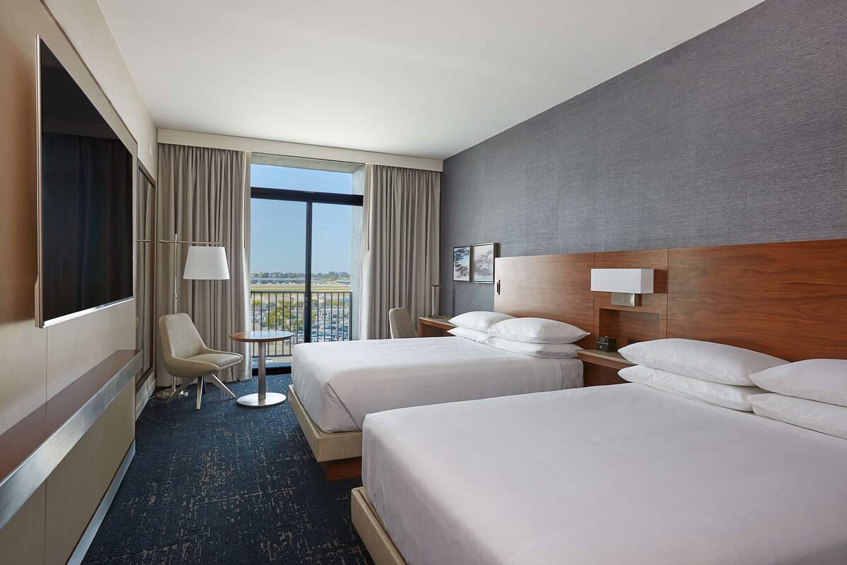 Ein Hotelzimmer mit zwei Betten, einem Schreibtisch, einem Stuhl und einem großen Fenster mit Blick auf die Stadt.
