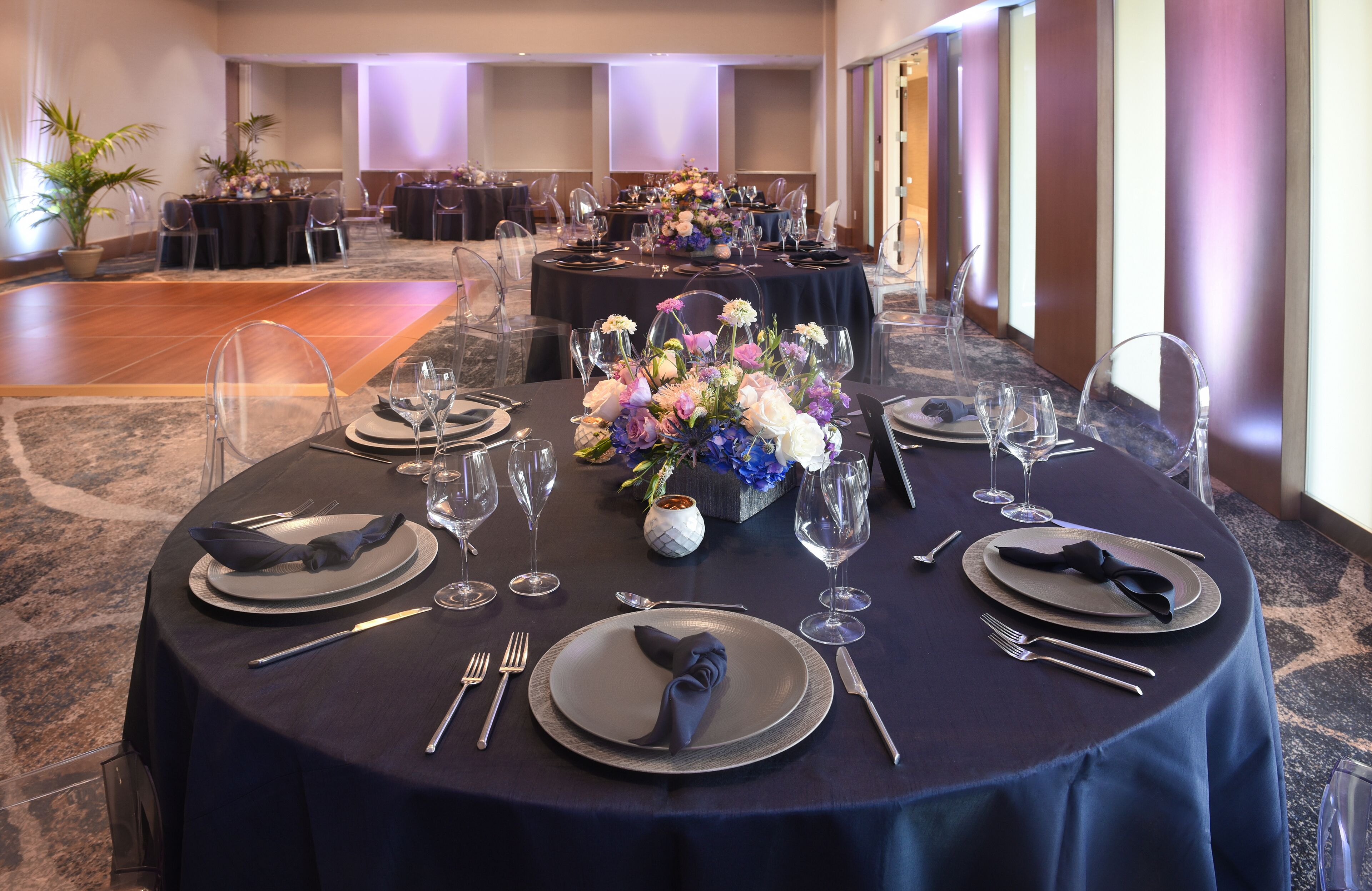 A banquet hall with round tables set for a formal event, featuring clear chairs, dark tablecloths, and floral centerpieces.
