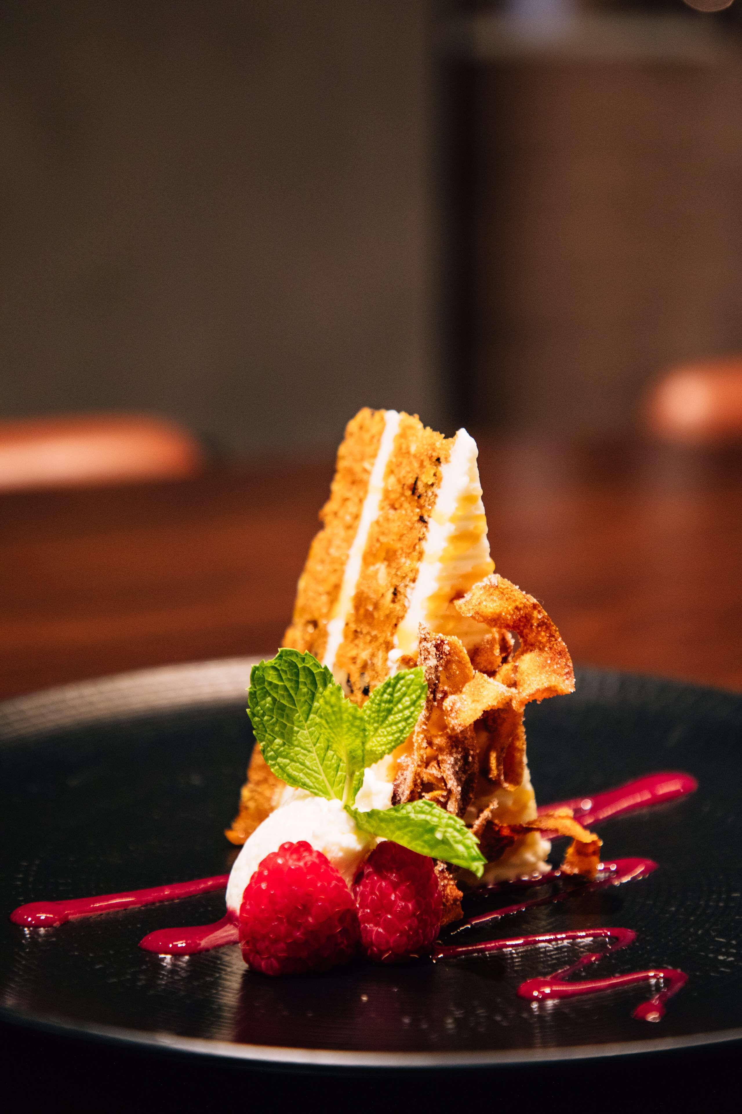 A dessert with a slice of cake, whipped cream, raspberries, and mint leaves on a black plate.