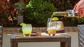 A tropical outdoor setting with a wicker table, a glass of beer, and a bottle of Corona Extra being poured. - Hyatt Regency Newport Beach West (Newport Beach)