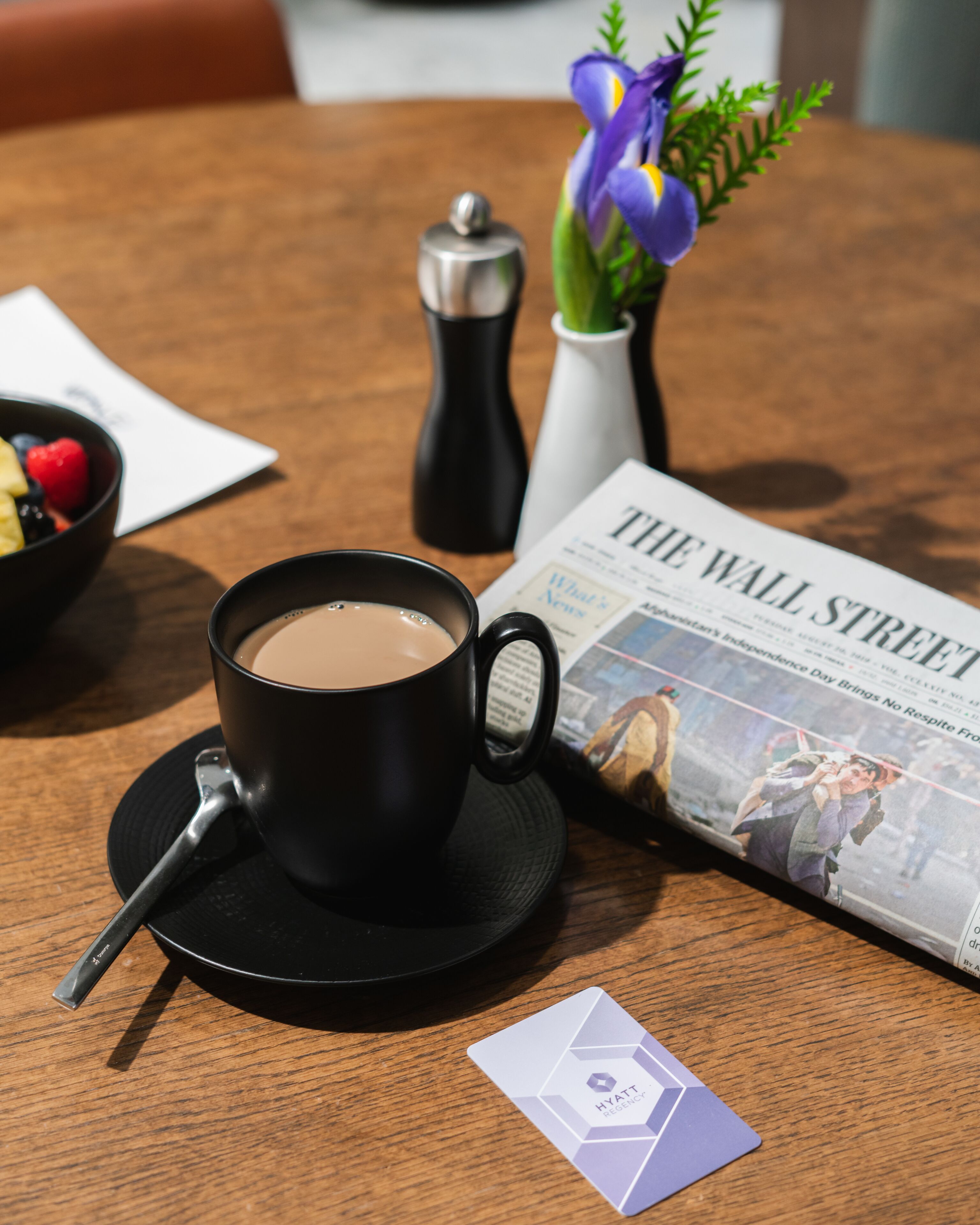 Une tasse de café posée sur une soucoupe, un salier, un vase avec des fleurs, un journal et une carte de visite, le tout sur une table en bois.