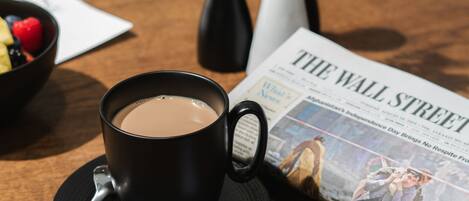 A cup of coffee on a saucer, a salt shaker, a vase with flowers, a newspaper, and a business card on a wooden table.