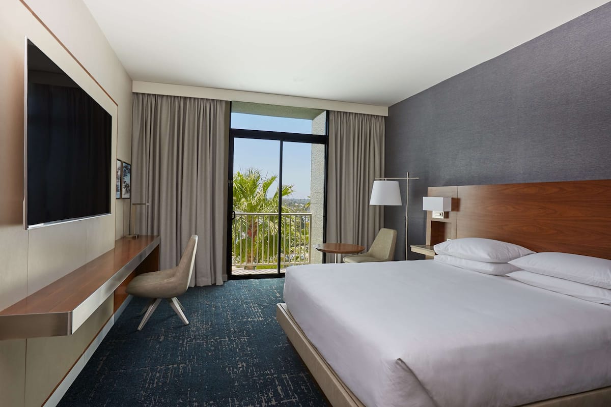 Ein Hotelzimmer mit einem großen Bett, einem an der Wand befestigten Fernseher, einem Schreibtisch mit Stuhl, einem kleinen Tisch mit Lampe und einem Balkon mit Blick auf Grünflächen.