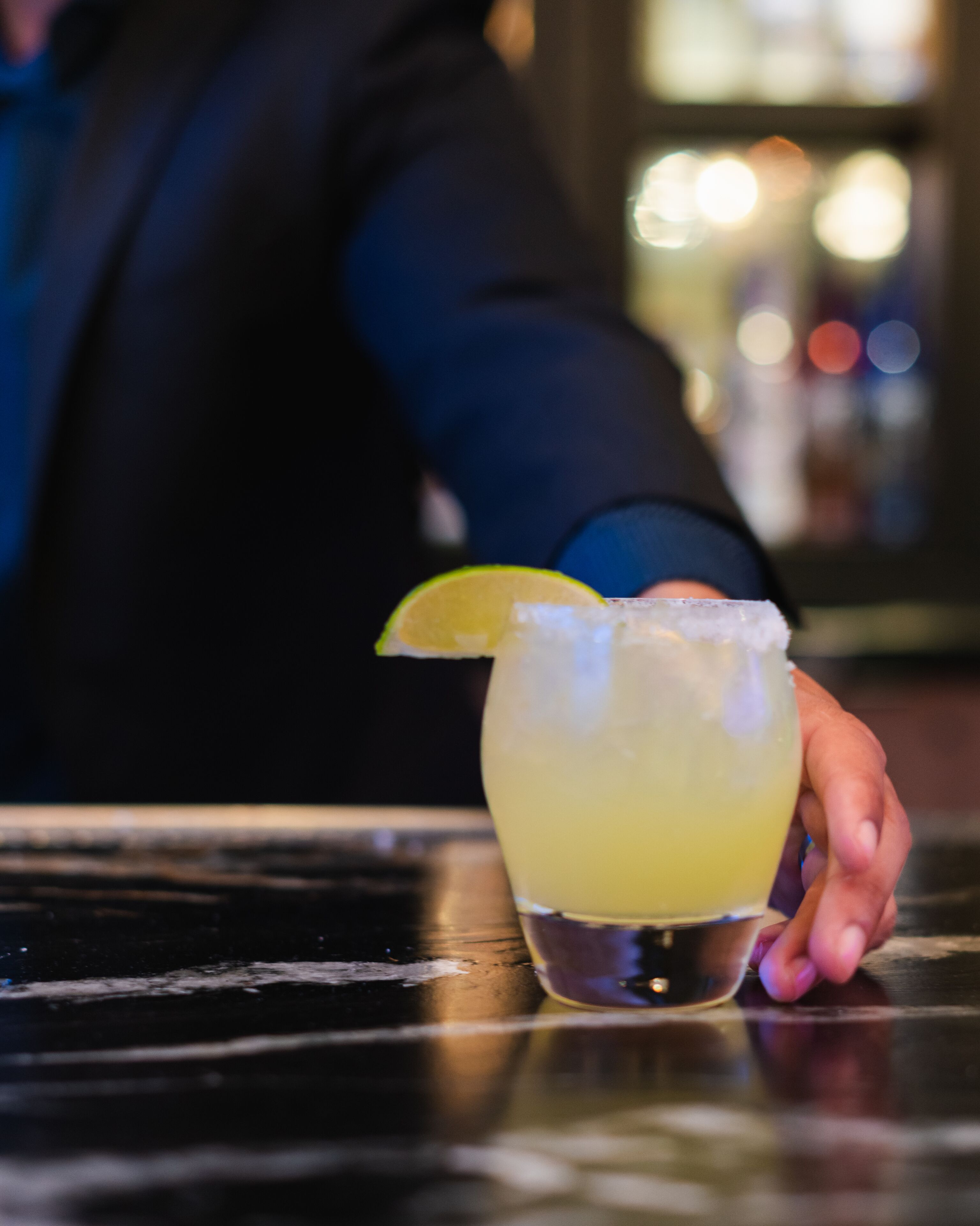 A hand holding a glass with a lime wedge, placed on a reflective surface.