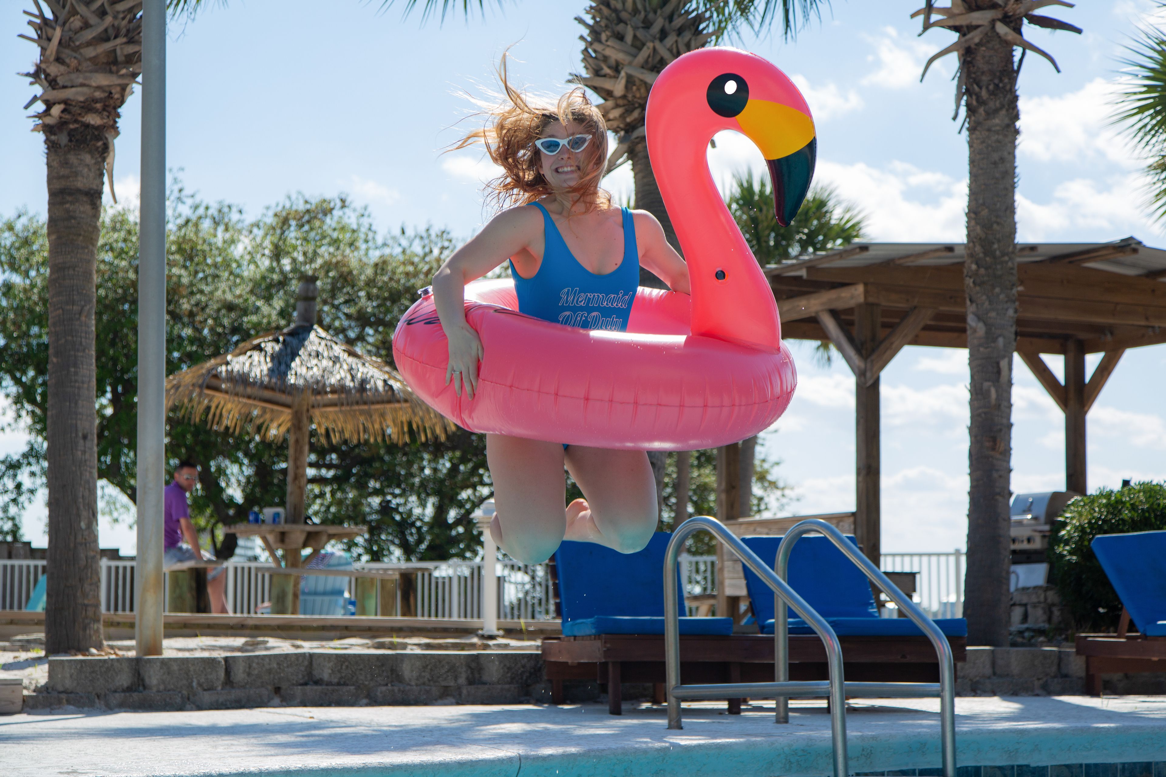 A person in a blue swimsuit with a mermaid design, standing on a pink flamingo float in a pool area with palm trees and lounge chairs.