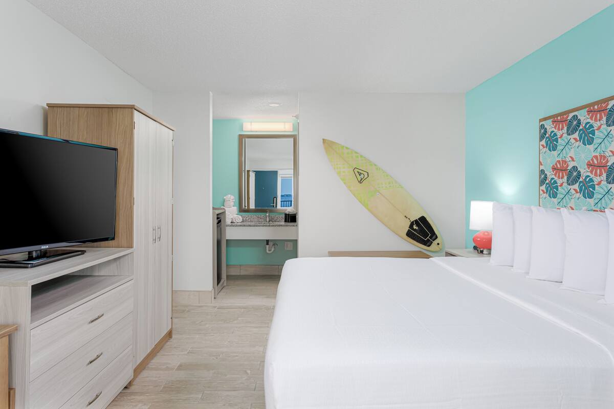 Un dormitorio con cama, televisión, armario y baño visible a través de una puerta abierta, además de una tabla de surf apoyada contra la pared.