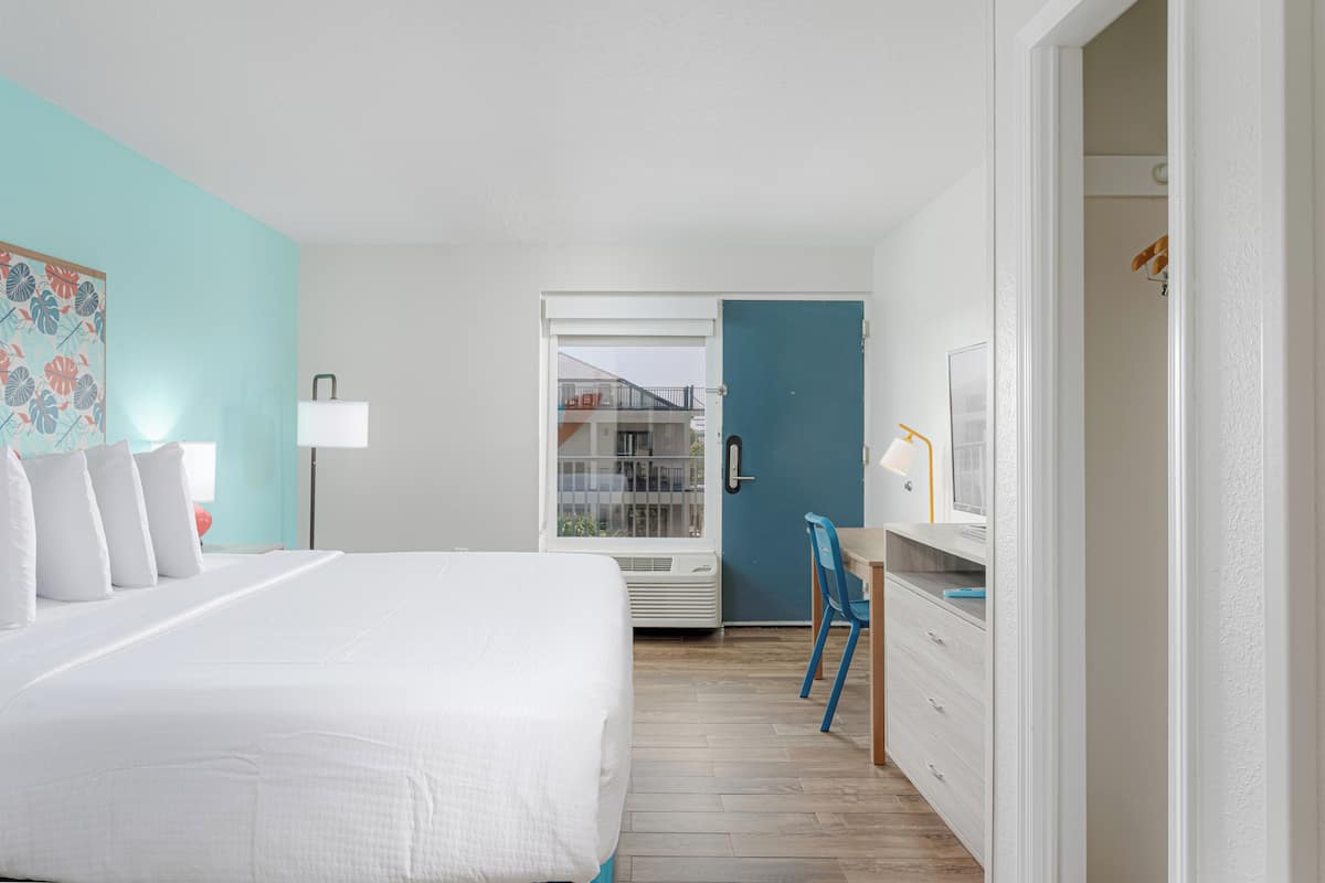 Habitación de hotel con cama, escritorio con silla, armario y puerta que da a un balcón.
