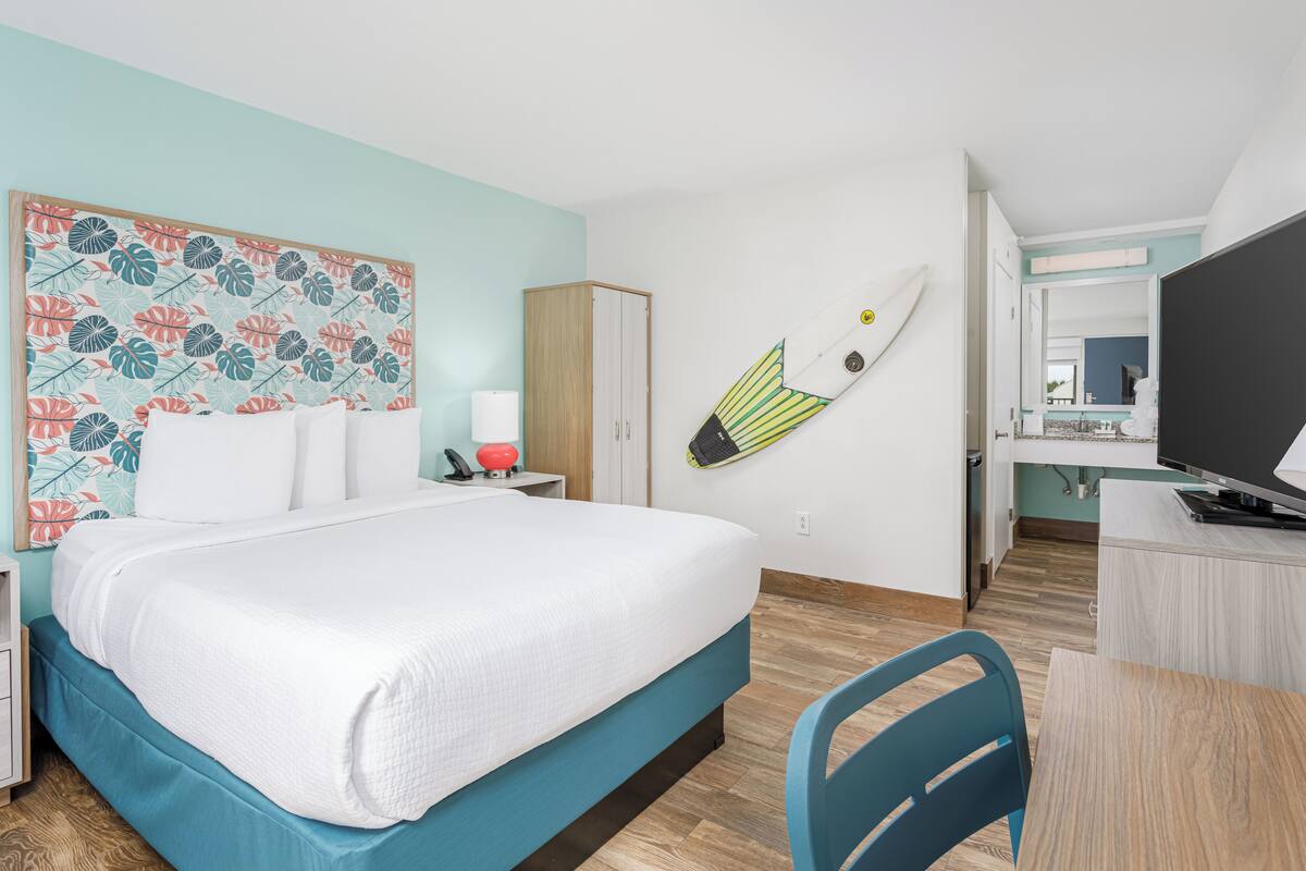 Habitación de hotel con cama, escritorio, silla, televisión y una tabla de surf colgada en la pared.