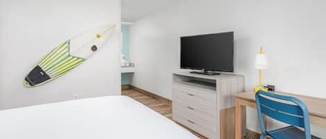 A hotel room with a bed, a desk with a chair, a television, and a surfboard on the wall.