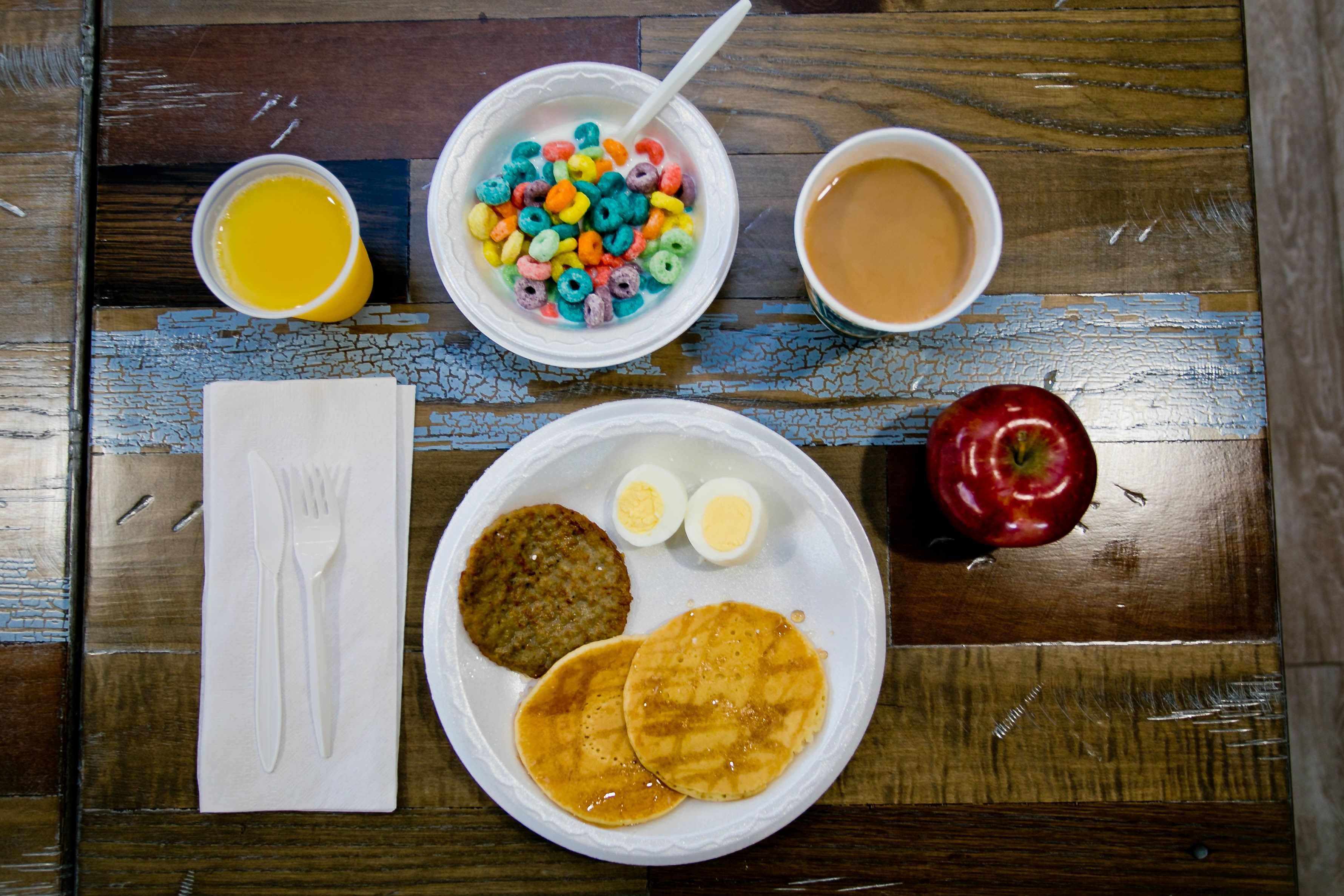 A meal with pancakes, eggs, cereal, and a red apple on a wooden table.
