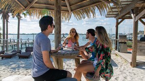 A wooden table with four people seated, overlooking a marina with boats and a sandy beach.