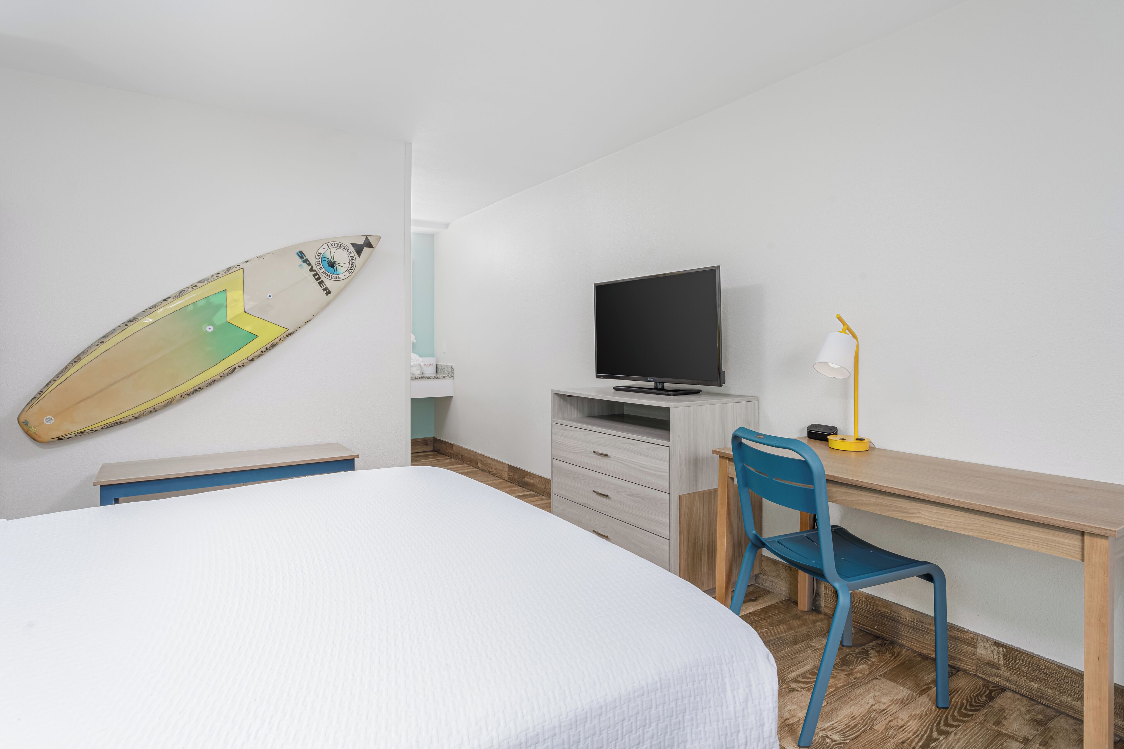 A bedroom with a bed, a desk with a chair, a television, and a surfboard on the wall.