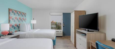 Habitación de hotel con dos camas, un escritorio con televisor, una silla de escritorio, una lámpara y una obra de arte floral grande en la pared.