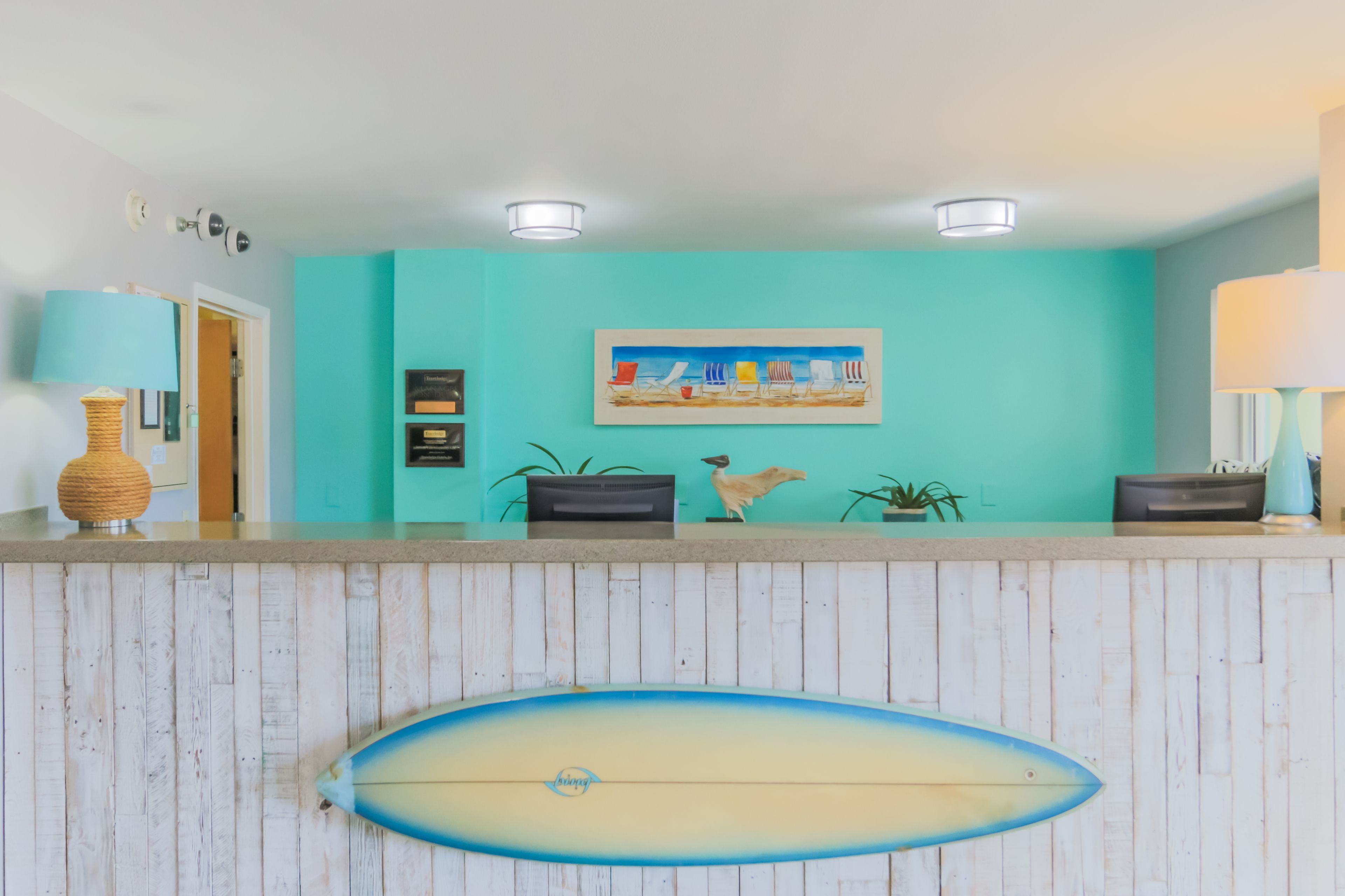 Un área de recepción con pared color turquesa, una decoración con tabla de surf, un cuadro de sillas de playa y un diseño de paneles de madera.