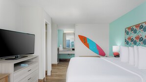 Habitación de hotel con cama, televisor sobre un aparador de madera, baño con espejo y una tabla de surf en la pared.