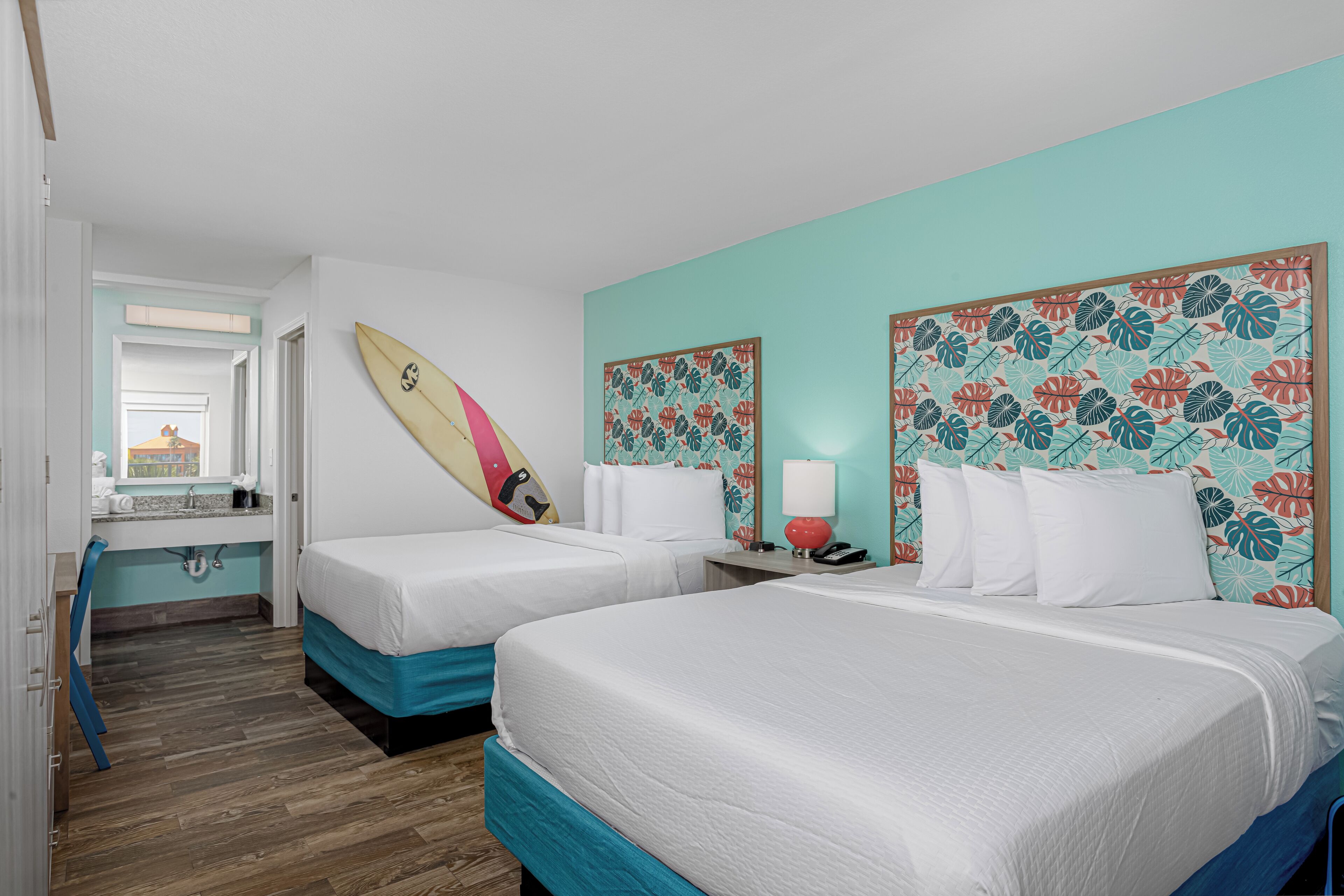 A hotel room with two beds, a surfboard, a bathroom with a sink, and a mirror.