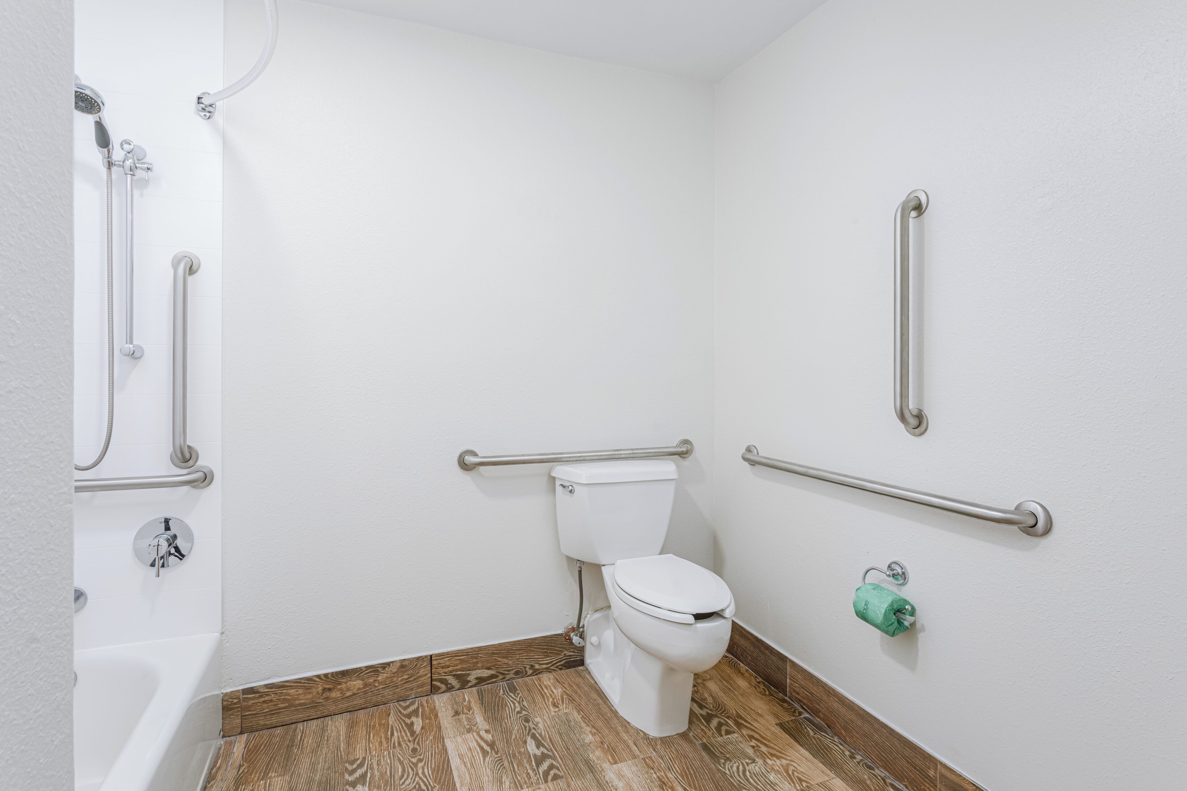 A bathroom with a white toilet, grab bars, a shower, and a wooden floor.