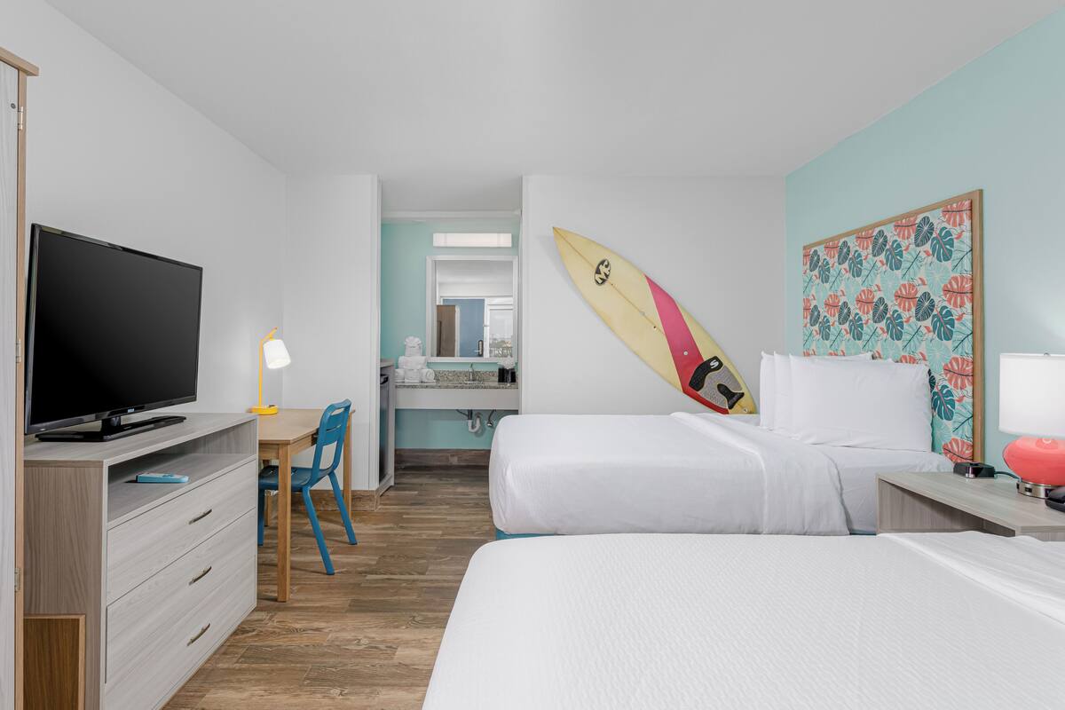 Una habitación de hotel con televisión, escritorio, silla, cama y una tabla de surf en la pared.