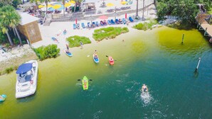 Vista aérea de una zona turística con playa, tablas de remo, kayaks y un barco amarrado cerca de la orilla.
