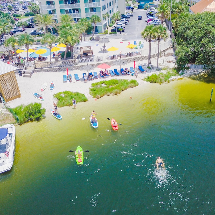 An aerial view of a resort area with a beach, paddleboards, kayaks, and a boat docked near the shore.