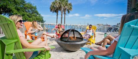 A group of people sitting on beach chairs around a fire pit on a sandy beach with palm trees and a clear sky in the background.