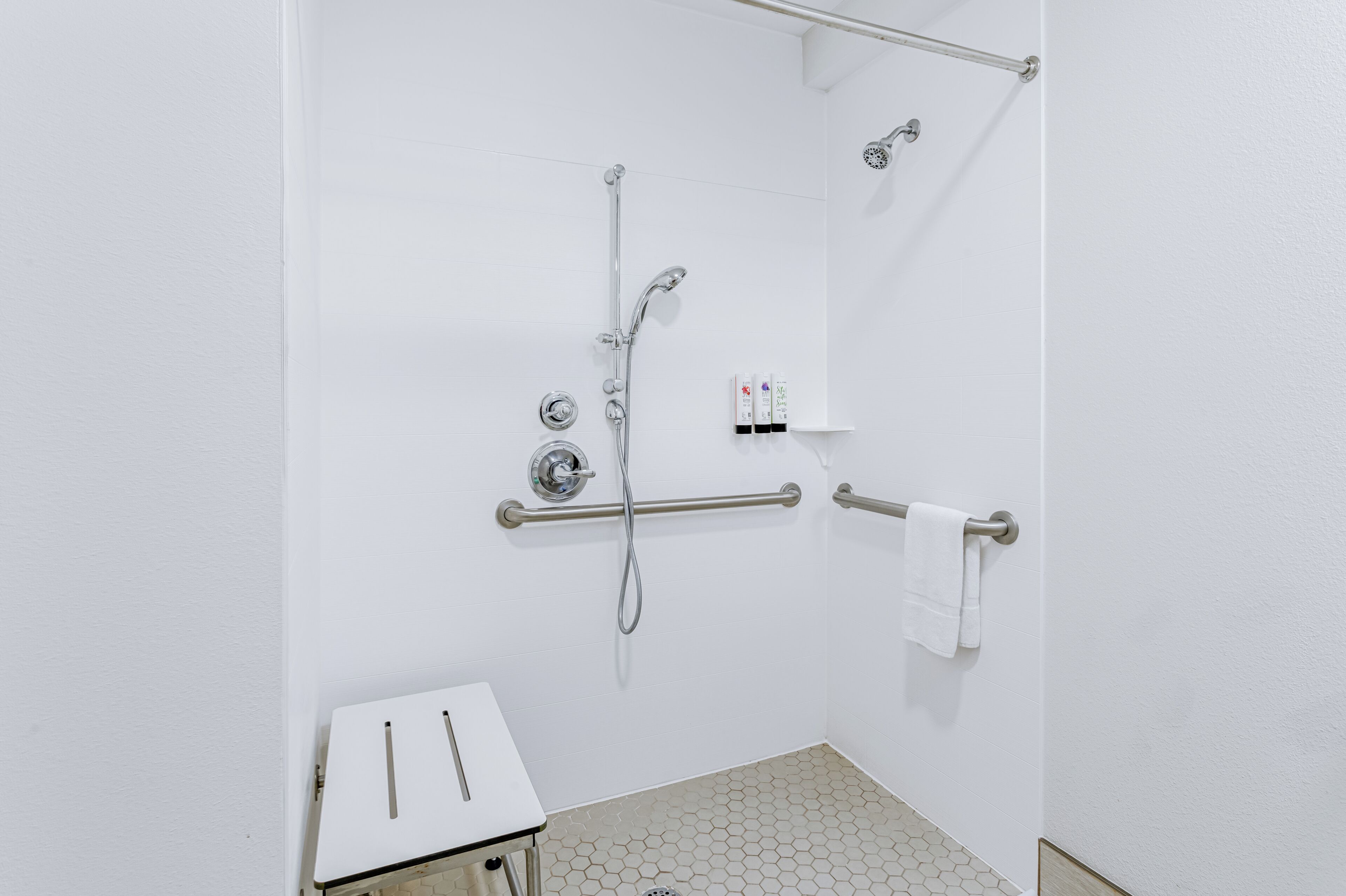 Une salle de bain blanche avec une douche, des barres d’appui, un porte-serviettes et une petite table sur laquelle est posée une serviette.