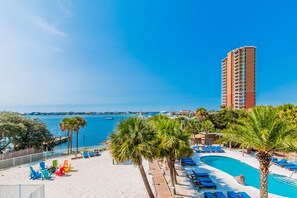 A resort with a pool, beach chairs, palm trees, and a view of the sea and boats.