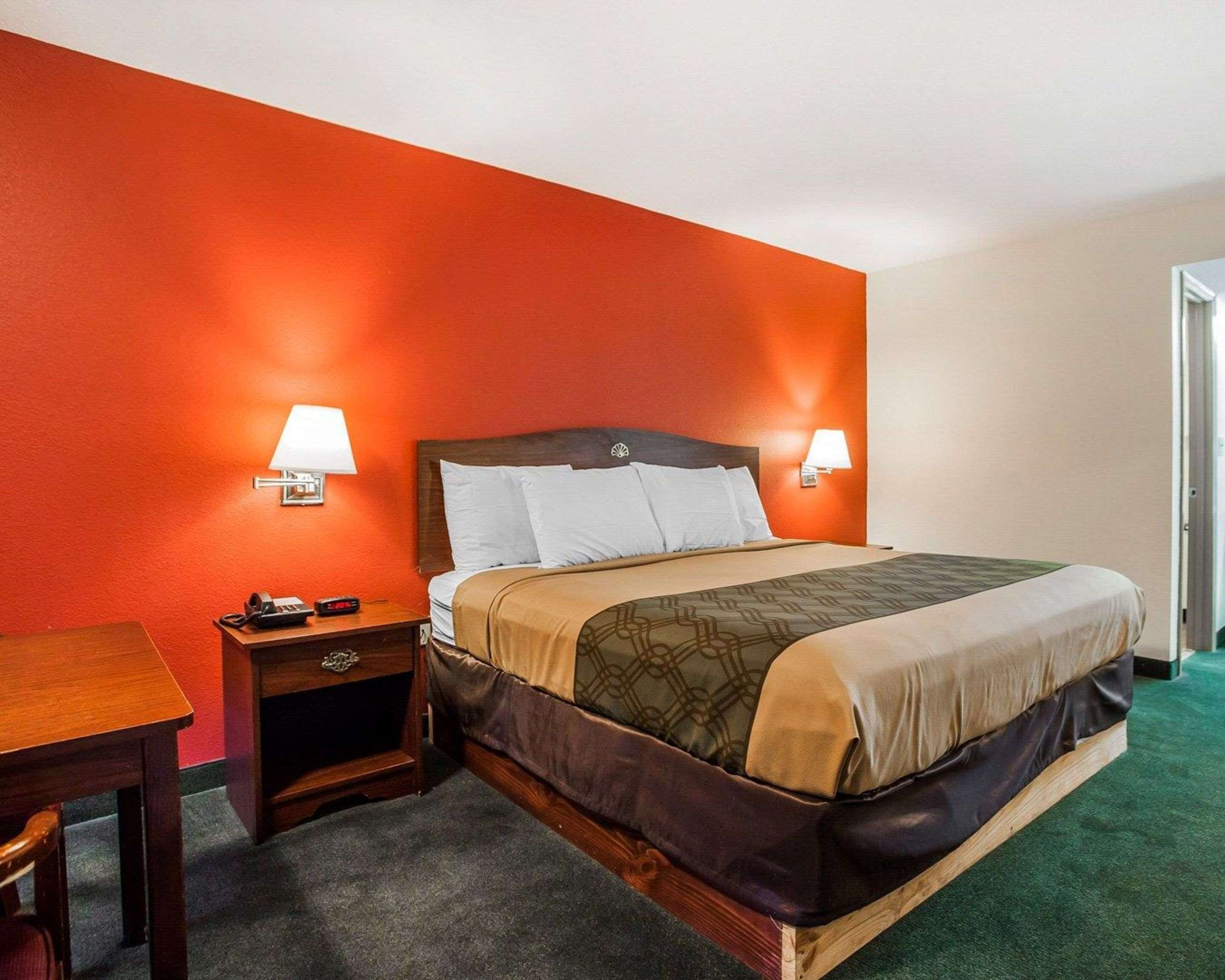 A hotel room with a single bed, two bedside lamps, a wooden nightstand with a telephone, and a desk with a chair.